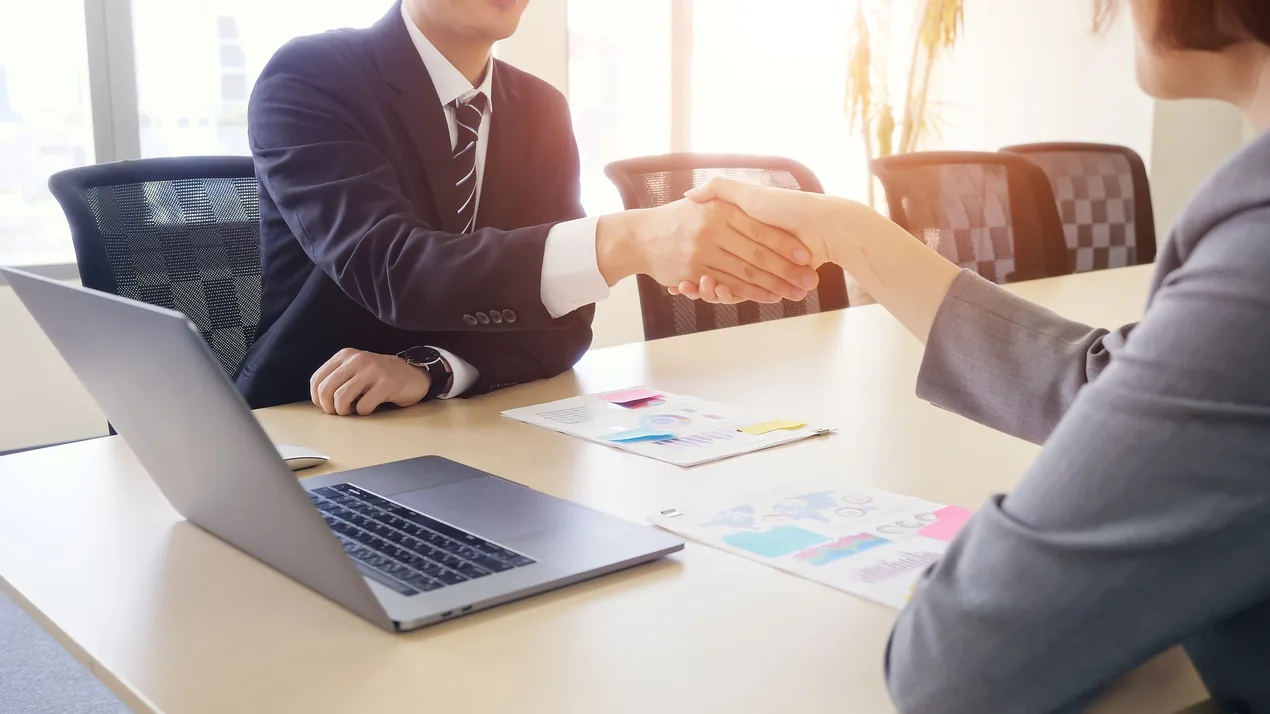 Two business professionals shaking hands across a desk with a laptop and paperwork