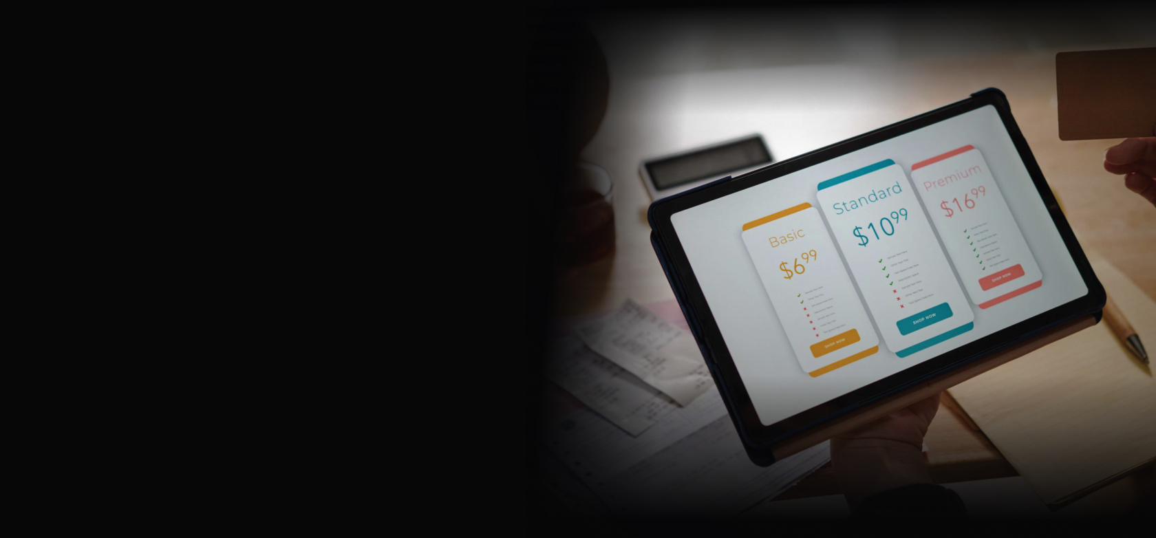 Hand holding a tablet showing three subscription pricing cards — Basic $6.99, Standard $10.99, and Premium $16.99 — on a desk with papers and a pen