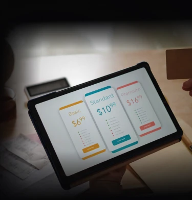 Hand holding a tablet showing three subscription pricing cards — Basic $6.99, Standard $10.99, and Premium $16.99 — on a desk with papers and a pen